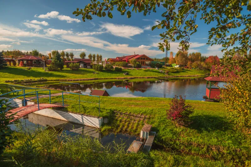 Słoneczny, jesienny widok na agroturystykę nad stawem. Na pierwszym planie, po lewej, mały drewniany mostek z niebieską barierką nad kanałem z betonowym przepustem. Obok, na trawiastym brzegu, krzew o czerwonych liściach. W centrum spokojny staw, w którym odbijają się budynki. Nad wodą po prawej czerwona altana. Brzegi porośnięte są zielonymi trawnikami i krzewami. W tle, na wzniesieniu, widać jasnodrewniany kompleks z czerwonymi dachami (pensjonat) i mniejsze domki letniskowe po lewej. Niebo jest błękitne z białymi chmurami. Z góry kadr zamykają gałęzie z liśćmi. Atmosfera jest spokojna i sielankowa.