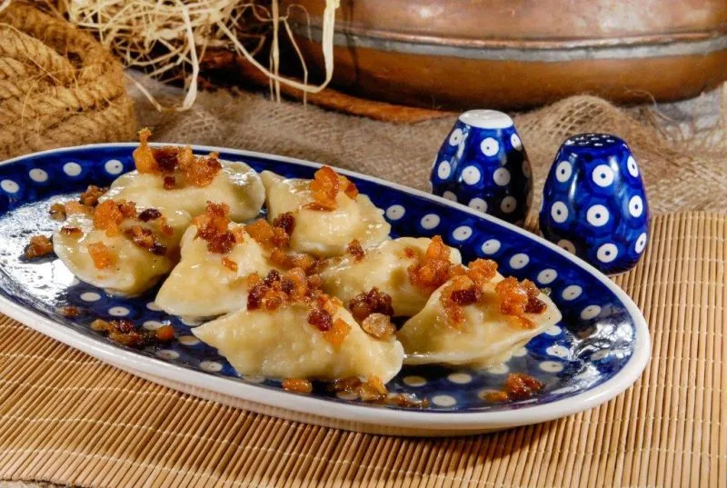 Pierogi z kaszą okraszone zarumienioną cebulką podane na zdobionym ceramicznym talerzu stojącym na drewnianym stole