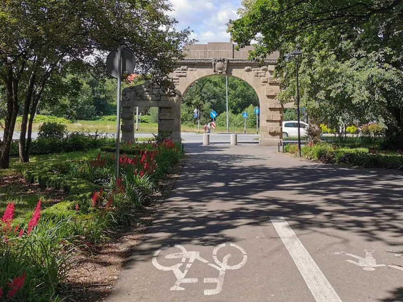 Zdjęcie przedstawia kamienną bramę oraz ścieżkę rowerową wysadzoną kwiatami