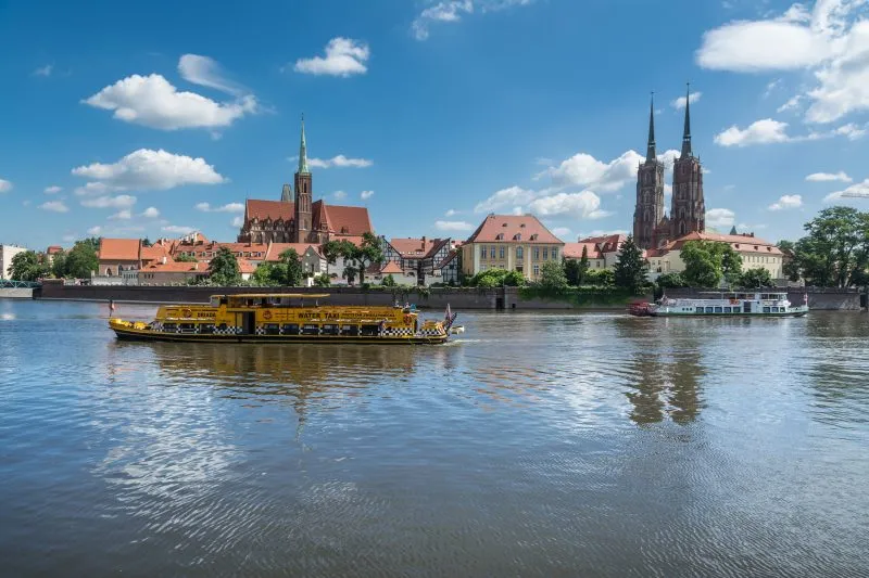Zdjęcie przedstawia rzekę z łodziami turystycznymi oraz zabytkowe zabudowania ostrowa tumskiego we wrocławiu w tle
