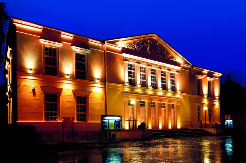 Zdjęcie przedstawia klasycystyczny budynek teatru w Jeleniej Górze ze sztucznym oświetleniem fasady na tle wieczornego nieba