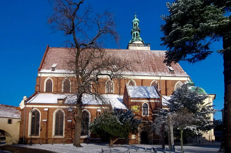 L'église gothique historique d'Oleśnica possède un chœur en briques, un haut toit rouge et une tour verte, entourés d'une cour enneigée.