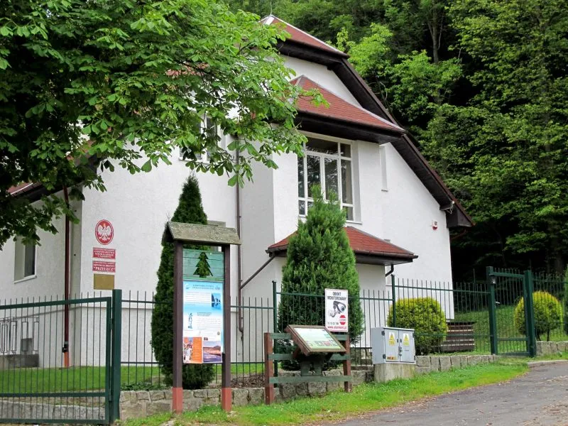 Na zdjęciu widać baił budynek zasłonięty z lewej strony koroną drzewa. Przed budynkiem stoi płot oraz dwie tablice informacyjne Karkonoskiego Parku Narodowego