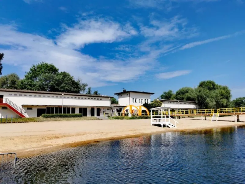 Na pierwszym planie zdjęcia widać kąpielisko łączące się z plażą. Na plaży znajduje się budka ratownika. Na drugim planie, w tle, znajdują się białe budynki kompleksu kąpieliska Kormoran