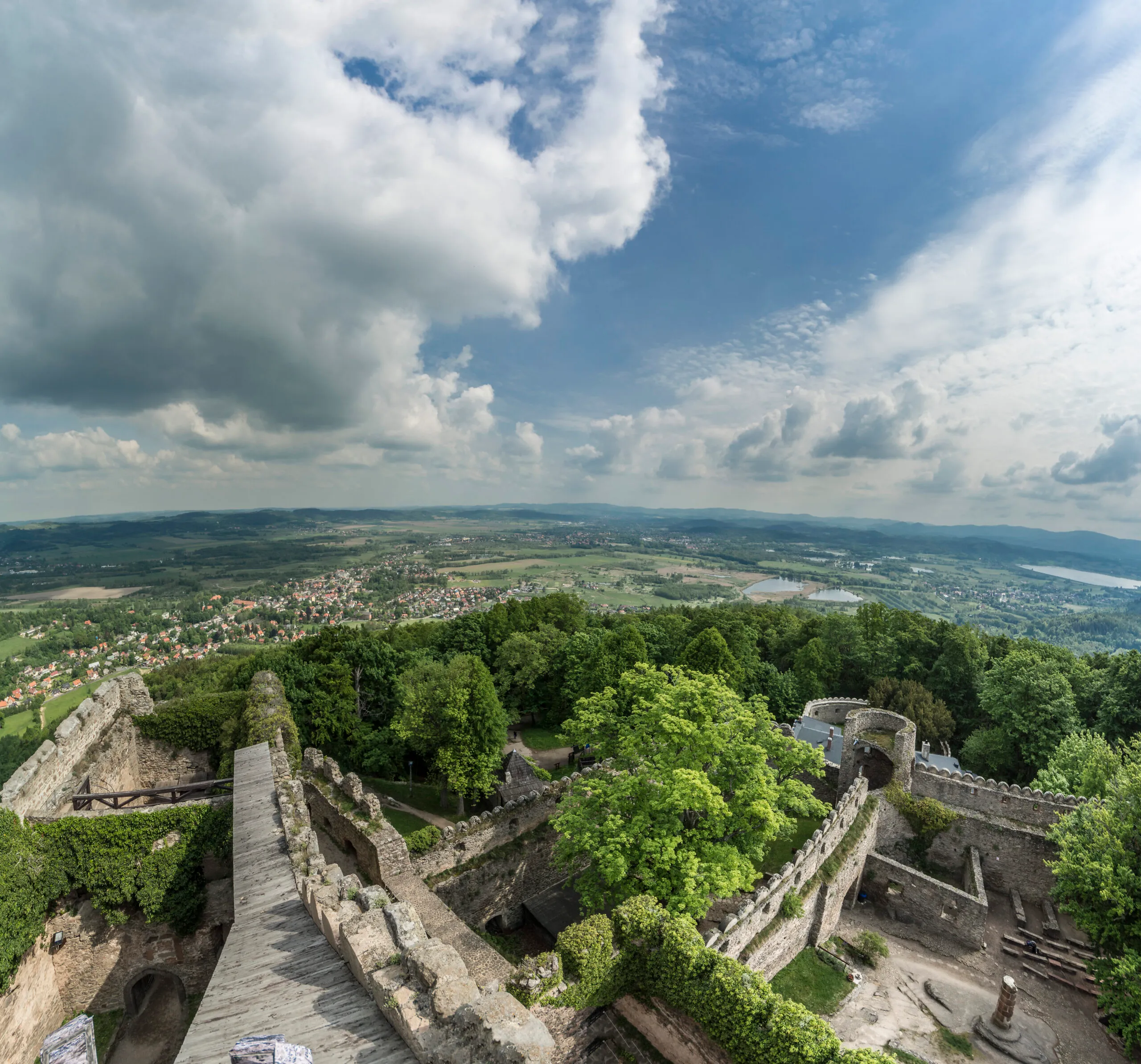 Widok znad murów kamiennego zamku otoczonego gęstym lasem. W tle ciekawa panorama najbliższej okolicy ze wzgórzami łąkami, zbiornikami wodnymi.