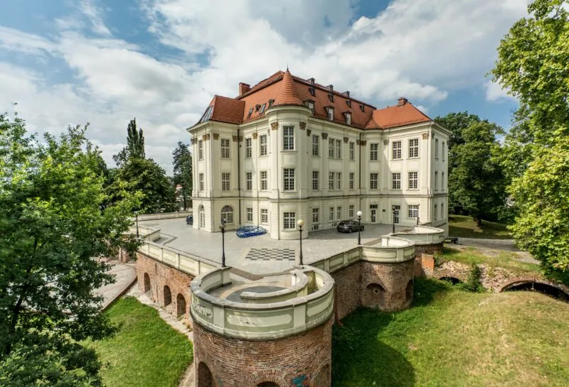 Zamek z czerwonym dachem, jasną elewacją oraz kwiatami na parapetach.