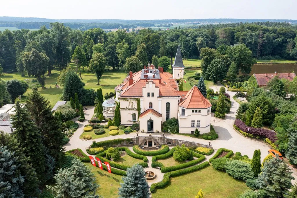 Widok na pałac z lotu ptaka, jasna elewacja, czerwony dach, przed pałacem piękny zadbany ogród.