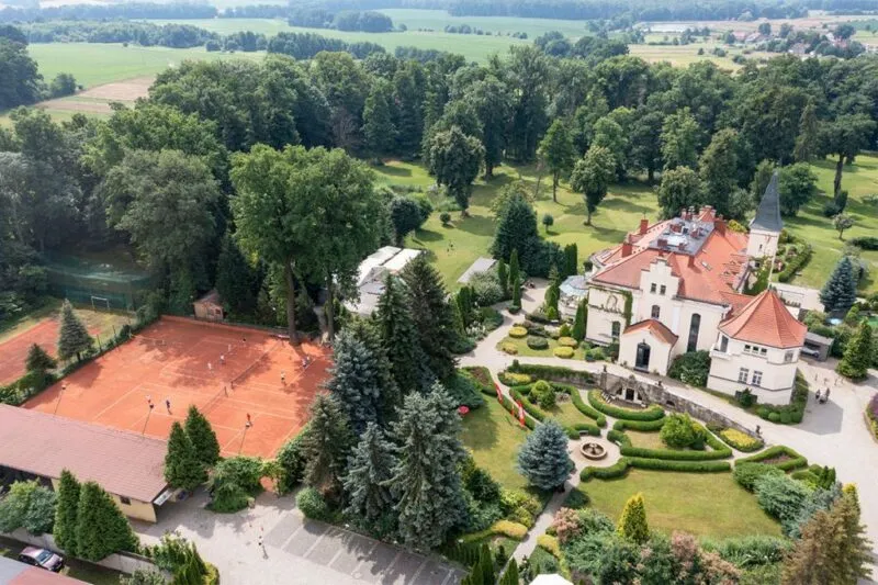 Widok na pałac z lotu ptaka, jasna elewacja, czerwony dach, przed pałacem piękny zadbany ogród. Obok pałacu kort tenisowy.