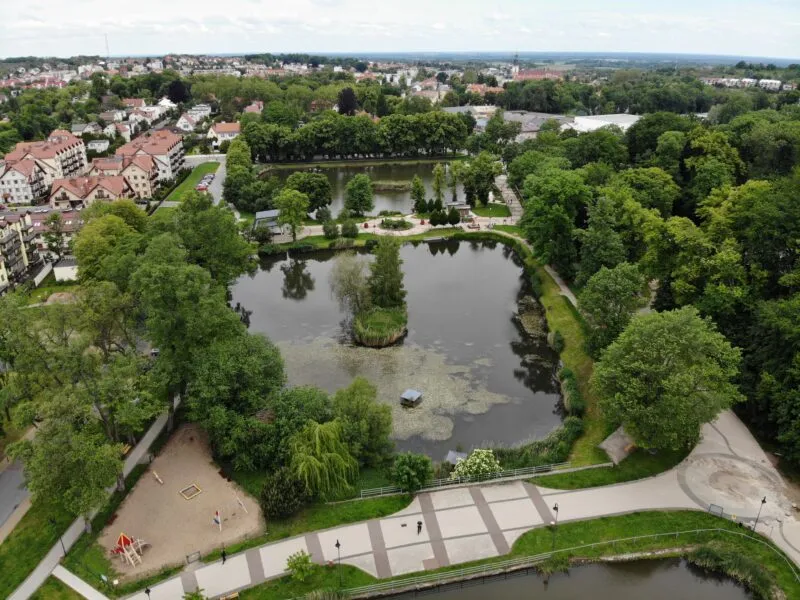 Głównym elementem zdjęcia są dwa stawy przedzielone alejką spacerową. Widać również liczne drzewa, a w oddali budynki.