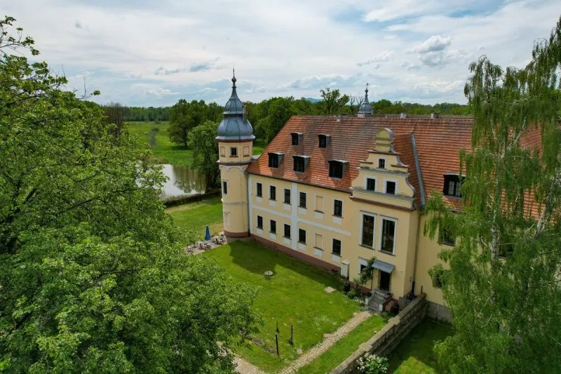 Il Palazzo di Krobielowice, con la sua facciata chiara, il tetto rosso e la torre d'angolo, sorge sopra uno stagno, circondato da un parco verde e alberi alti.