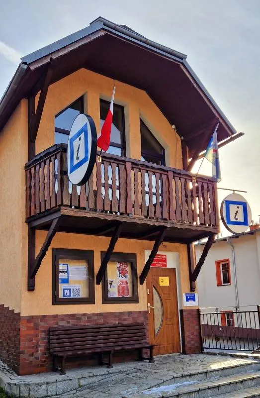 Wąski piętrowy budynek z nieproporcjonalnie dużym balkonem. Na budynku widać podwójne oznaczenie punktu informacji turystycznej oraz dwie flagi. Przy drzwiach wejściowych stoi ławka.