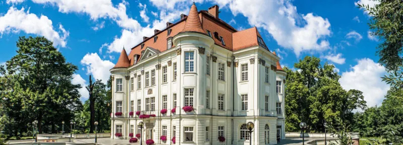 Zamek z czerwonym dachem, jasną elewacją oraz kwiatami na parapetach.