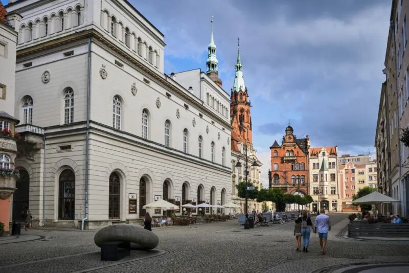 Wysoki budynek teatru w Legnicy z kostkową płytą Rynku, w tle ceglane wieże gotyckie katedry