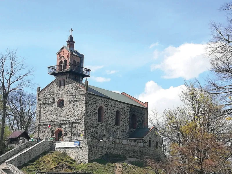 Widok na kamienny, nieotynkowany kościół z dwuspadowym dachem i niewielką wieżą w centralnej części fasady na tle nieba.