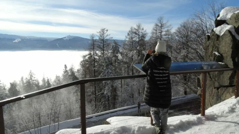 Zimowy widok z niezadaszonego tarasu widokowego znajdującego się na skraju góry z widocznymi w oddali Karkonoszami. Na pierwszym planie fotografująca widok turystka.