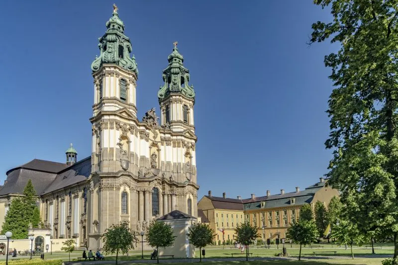 Dwie wysokie wieże kościelne w stylu barokowym, obok zabytkowe żółte budynki opactwa