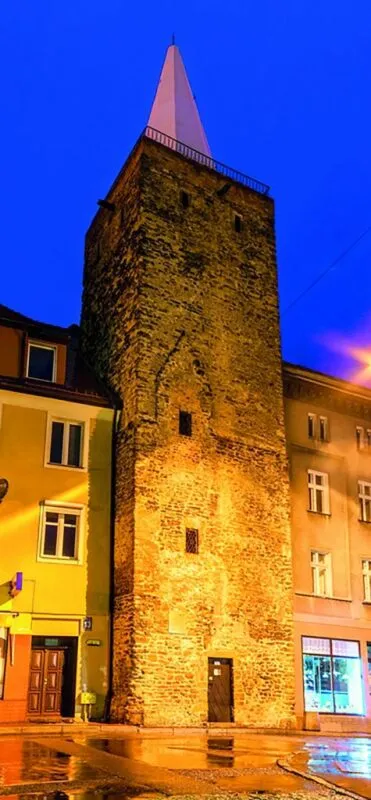 Nocny widok na średniowieczną, kamienną wieżę, kwadratową w swym planie, z przylegającymi do niej budynkami mieszkalnymi.