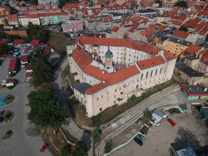 Zdjęcie bryły zamku, wykonane z drona. Widoczny jest niemalże trójkątny rzut obwarowań, łososiowy kolor fasady oraz czerwona dachówka.