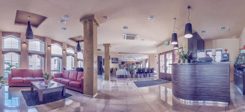 Recepcja hotelowa wraz z pomieszczeniem ogólnodostępnym. Na zdjęciu widać kanapy do siedzenia kolumny w pomieszczeniu oraz duże okna wpuszczające wiele światła do środka