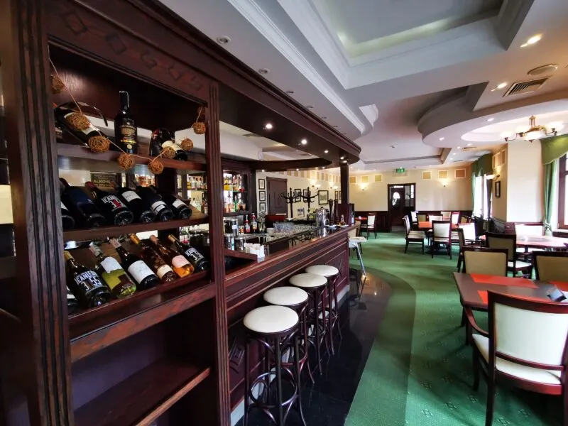Il bar dell'hotel in legno scuro presenta scaffali con bottiglie, sgabelli alti e file di tavoli nell'accogliente interno dell'Hotel Bukowy Park.