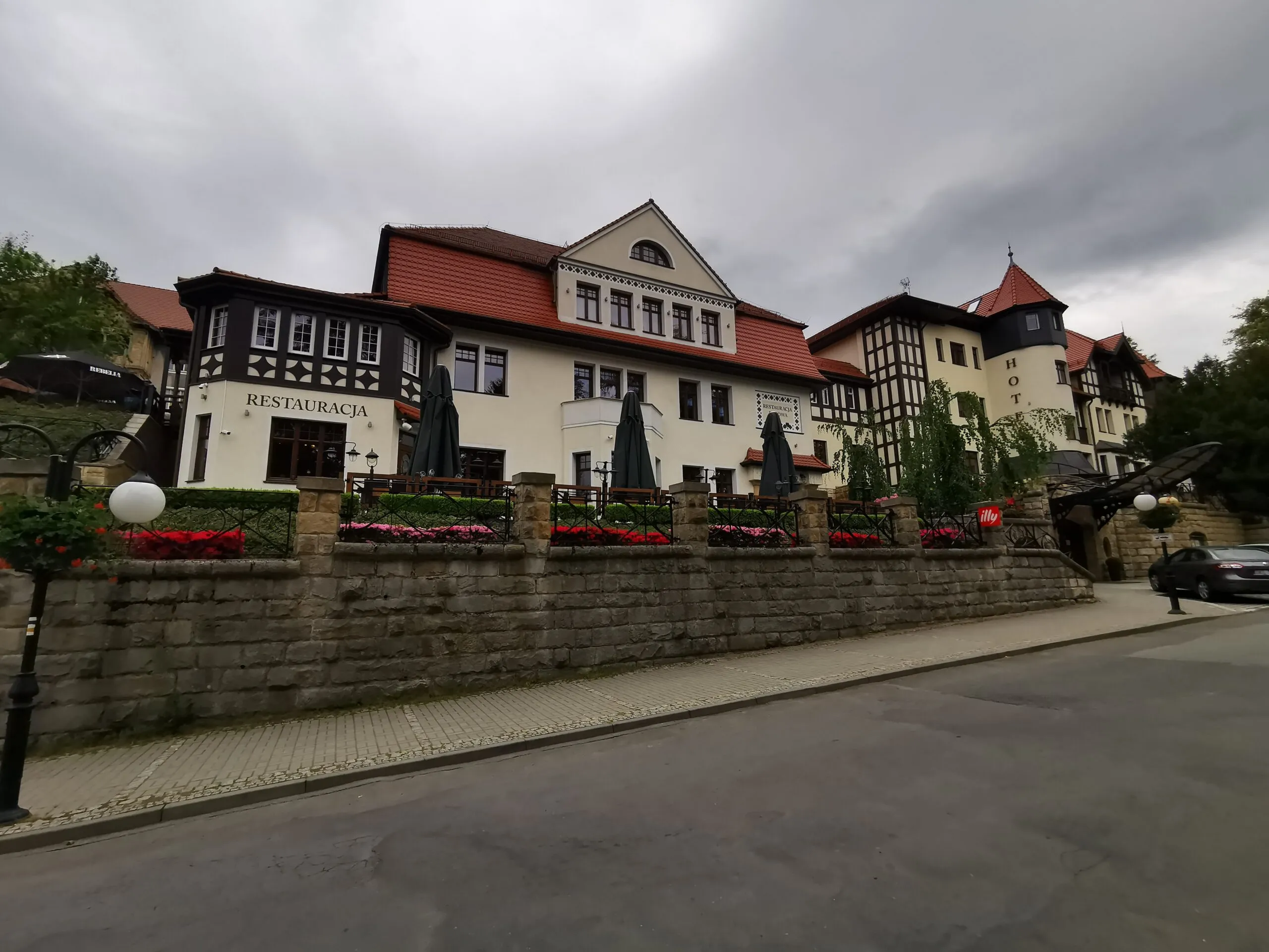 The bright building of the Bukowy Park Hotel in Polanica-Zdrój with dark wooden gables and a red roof stands by the street behind a stone wall.