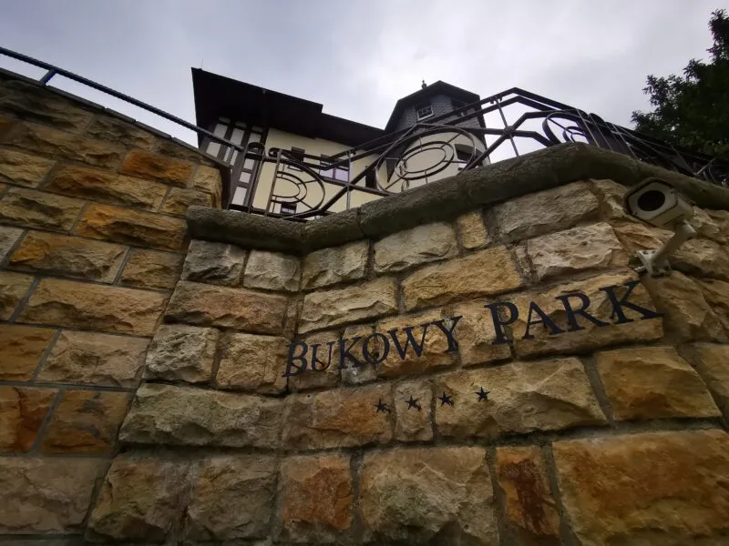 Il muro di pietra presso l'Hotel Bukowy Park a Polanica-Zdrój è decorato con una balaustra in metallo e il nome della struttura inciso, visibile sullo sfondo di un cielo nuvoloso.