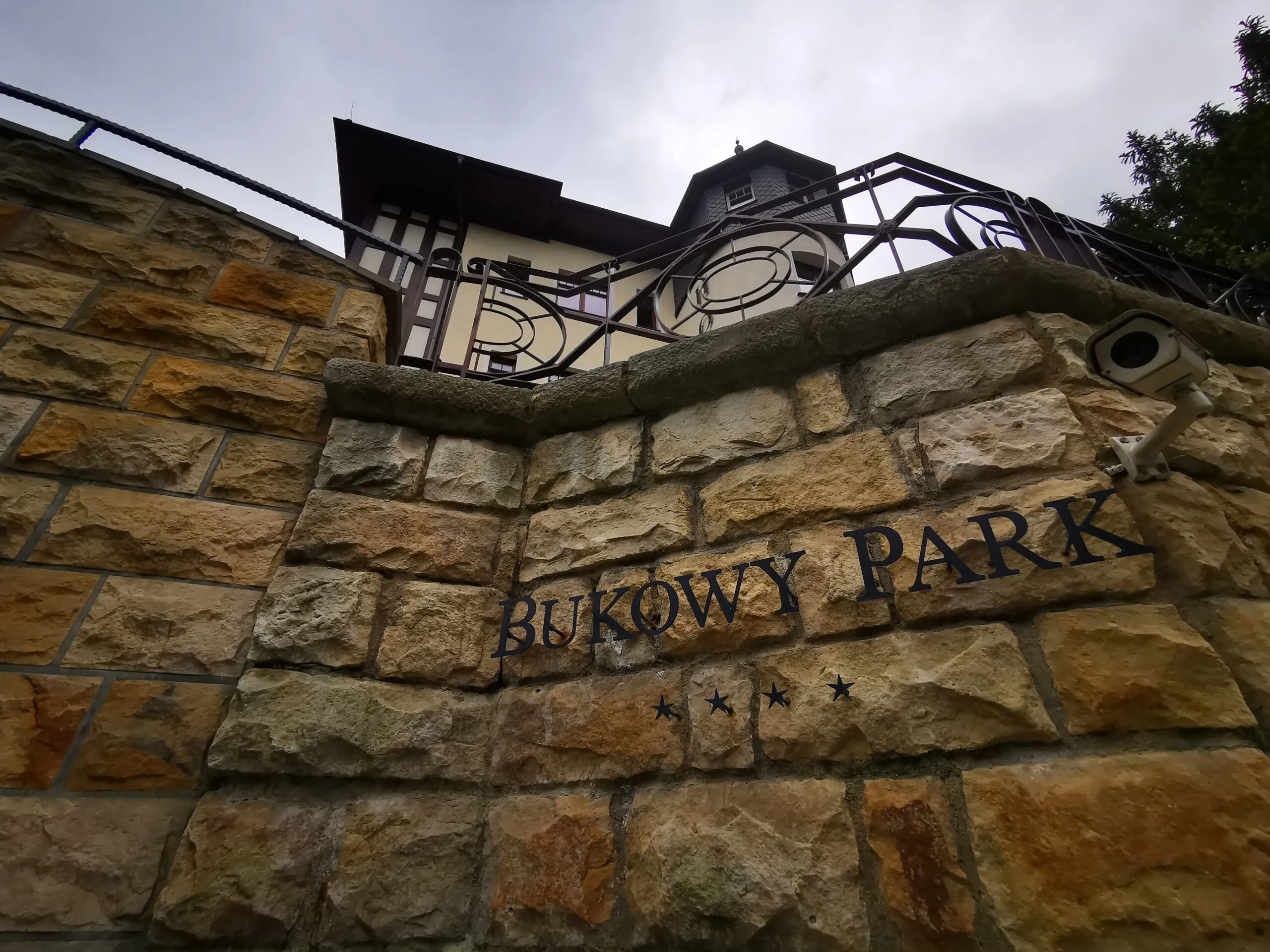 The stone wall by the Bukowy Park Hotel in Polanica-Zdrój is decorated with a metal railing and the engraved name of the facility, visible against a cloudy sky.