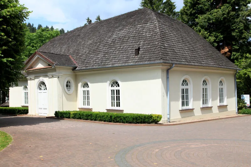 Skromny, mały beżowy budynek, z łukowymi oknami, pokryty czarną dachówką. 
