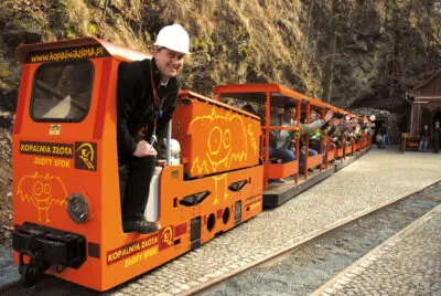 The orange tourist train of the Gold Mine in Złoty Stok waits for passengers in front of the rock entrance, with the train driver standing by the locomotive.