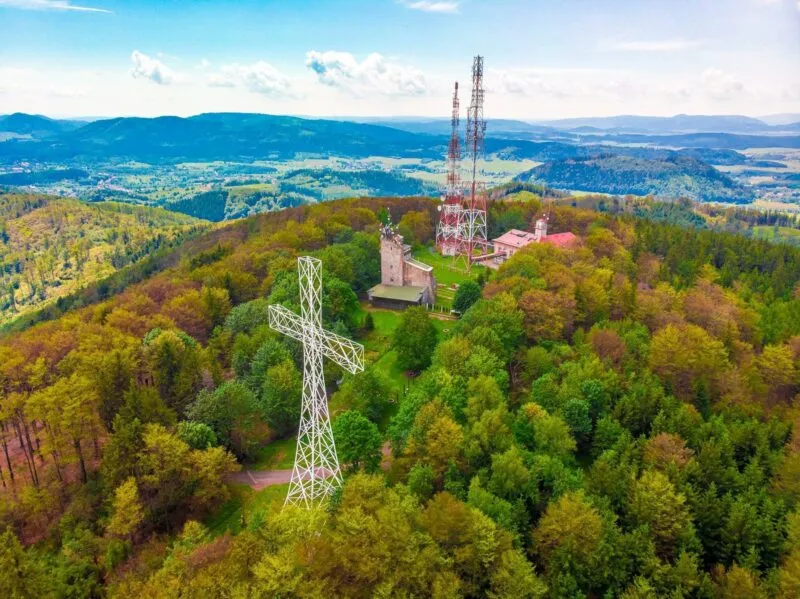 Metalowy krzyż oraz wieża radiowo-telewizyjna na szczycie góry, porośniętej zielonym lasem.
