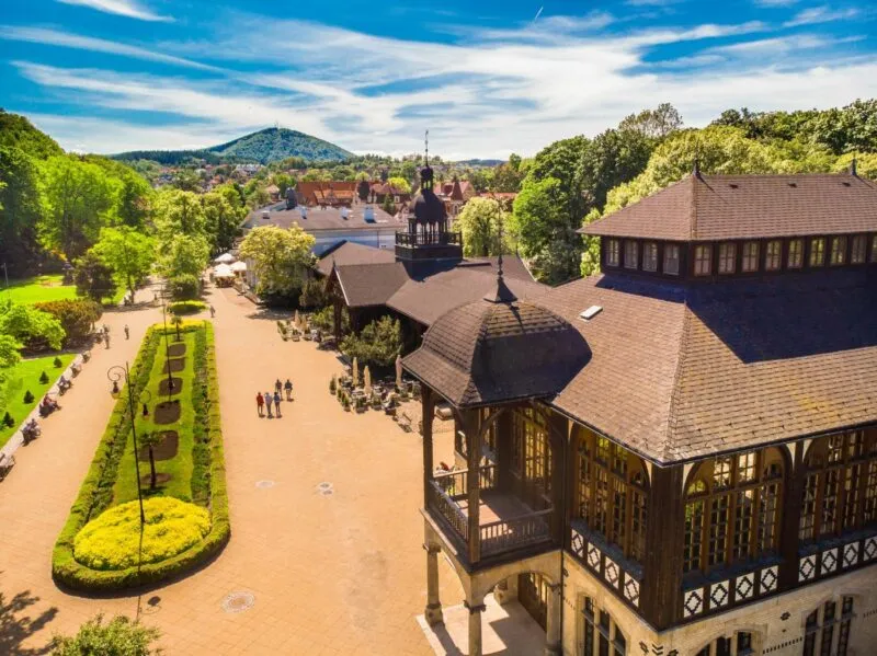 Zdjęcie z drona przedstawiające park zdrojowy latem wraz z drewnianym, bogato zdobionym budynkiem zabytkowej pijalni wody.