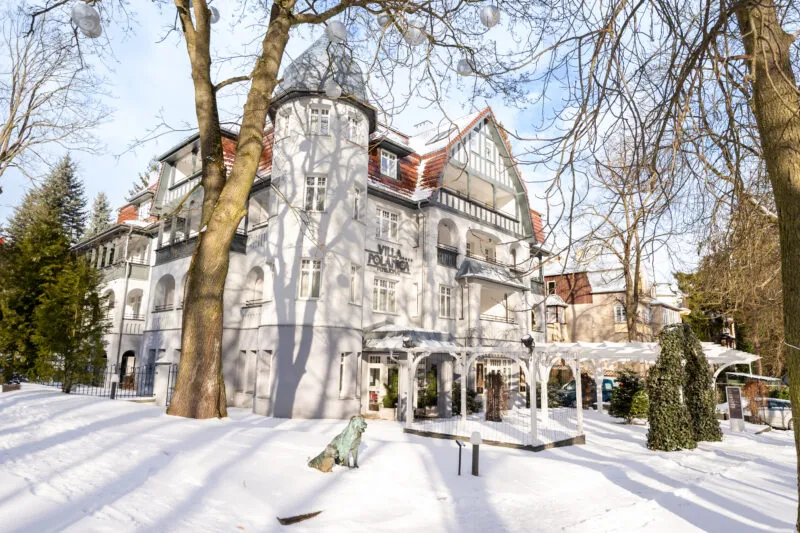 La facciata bianca di Villa Polanica con la sua torretta, i frontoni in legno e il tetto rosso si erge in un parco invernale tra alberi spogli e neve fresca.