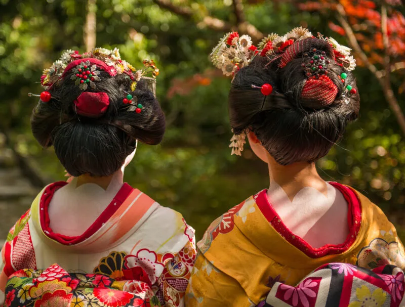 Deux femmes en kimonos japonais colorés se tiennent de dos sur un sentier forestier, présentant des coiffures élaborées ornées de fleurs et d'accessoires rouges.