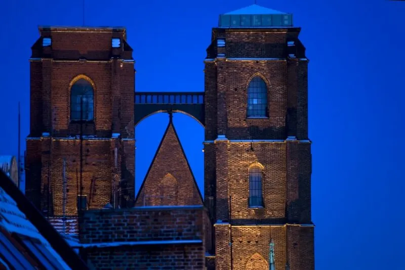 Widok na murowane, gotyckie wieże z widocznym mostkiem łączącym je na znacznej wysokości na tle wieczornego nieba