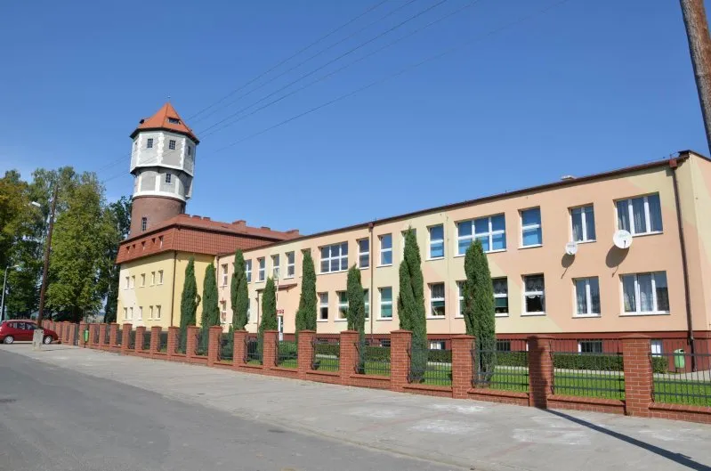 Ceglana wieża ciśnień z odciętym szczytem w kolorze grafitowym oraz sąsiadujący z nią podłużny budynek szkoły.