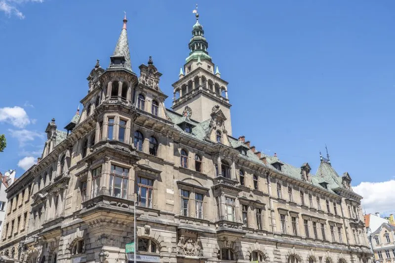 Widok na kłodzki ratusz, budowla w stylu neorenesansowym, wyróżniająca się smukłą wieżą zegarową oraz bogato zdobioną fasadą z detalami architektonicznymi.