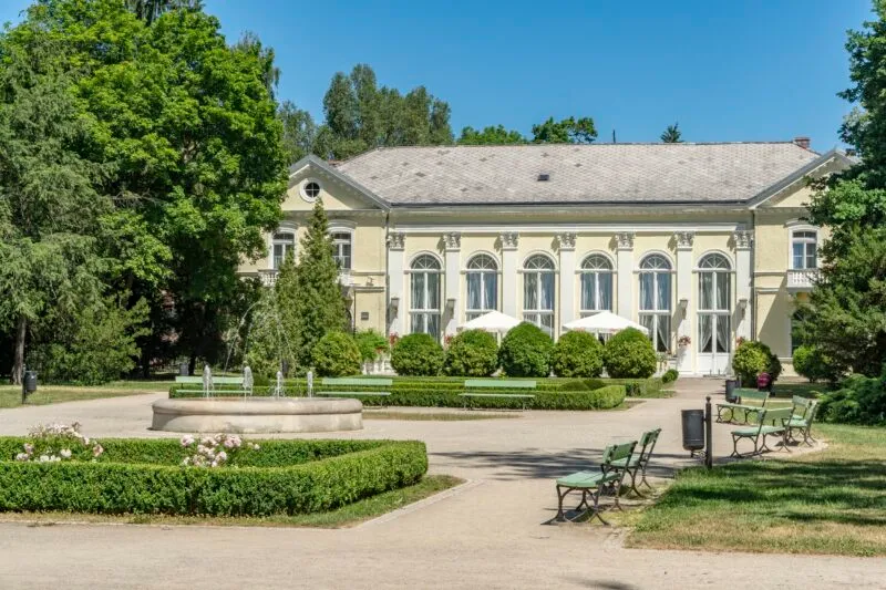Zabytkowy jasny pałac otoczony zielenią, na pierwszym planie okragła fontanna