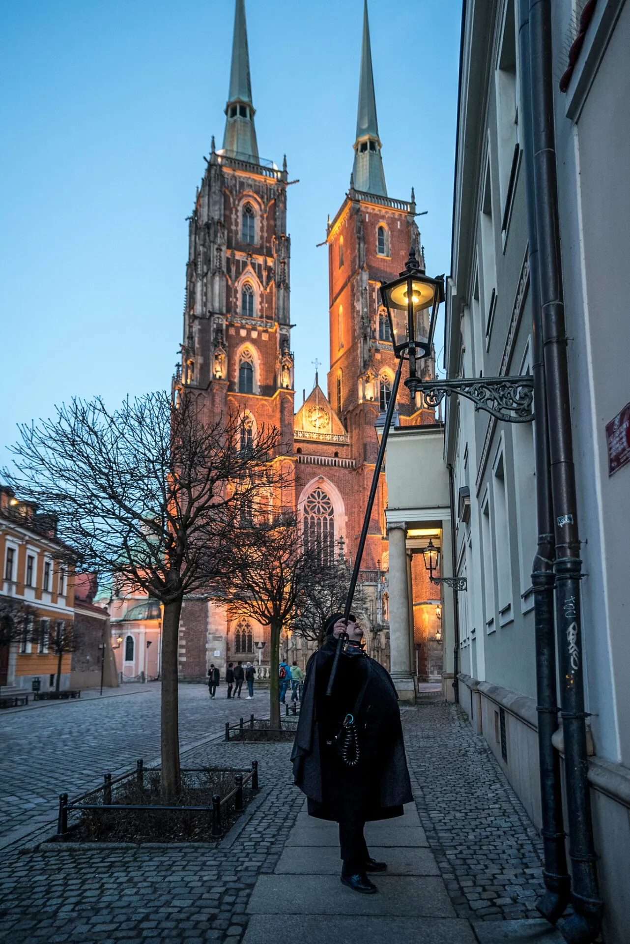 Widok na gotycką budowlę z cegły, wyróżniająca się strzelistymi wieżami, bogato zdobioną fasadą oraz misternymi witrażami w wysokich oknach. Na pierwszym planie latarnik zapalający latarnię,