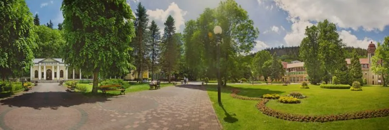 Widok na część uzdrowiska - park, w którym rosną drzewa, otoczone zadbanym trawnikiem, pośrodku ścieżka spacerowa oraz w tle widoczne budynki.