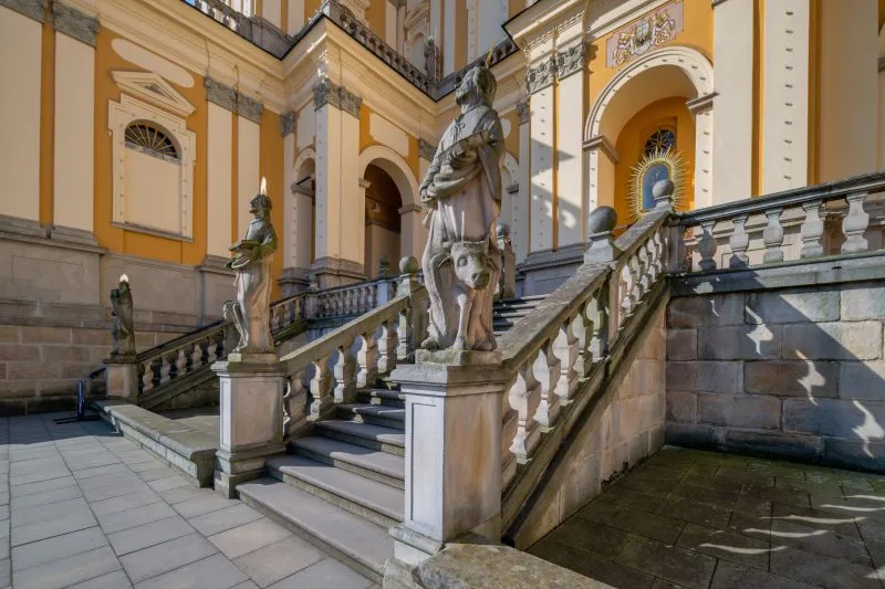 Na zdjęciu widać okazałe sanktuarium w Wambierzycach z monumentalnymi schodami prowadzącymi do barokowej fasady ozdobionej kolumnami i wieżami.