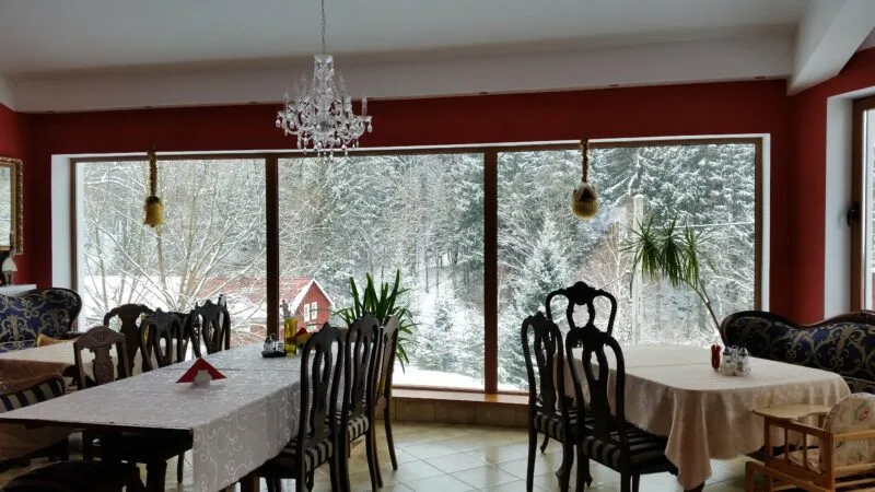 The bright dining room of the Rudy Guesthouse features tables with white tablecloths, wooden chairs, a crystal chandelier, and large windows overlooking a snow-covered garden.