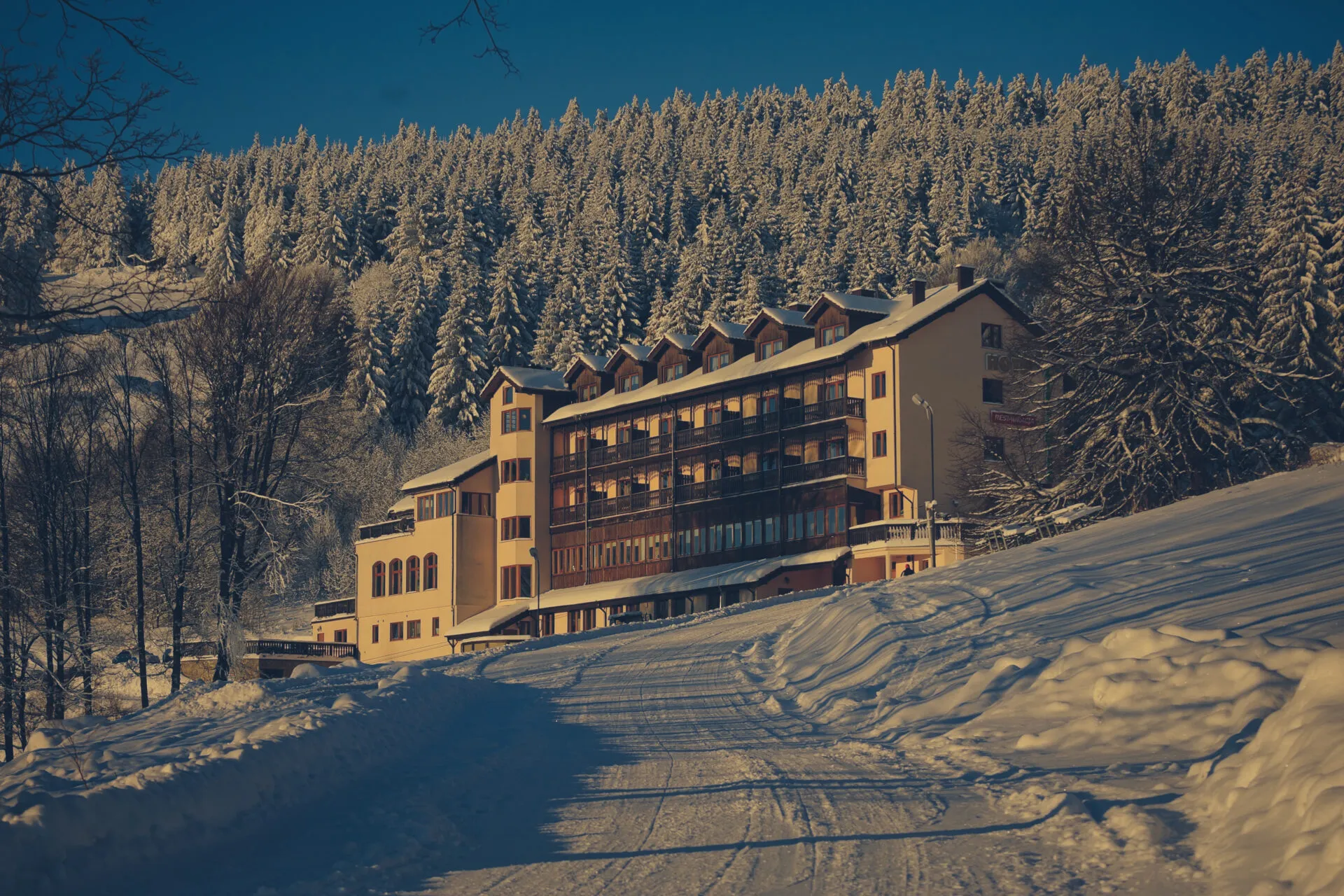 Duży, kilkupiętrowy budynek hotelu, położony na lekkim wzniesieniu, tuż przed lasem iglastym, skąpany w śniegu.