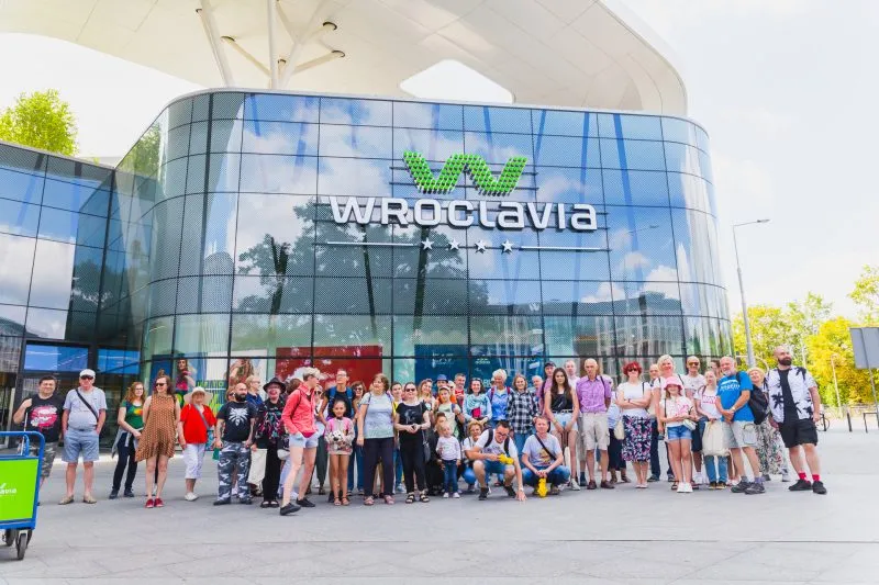 Grupa turystów pozuje do zdjęcia z wejściem do galerii handlowej Wroclavia widocznej w tle 