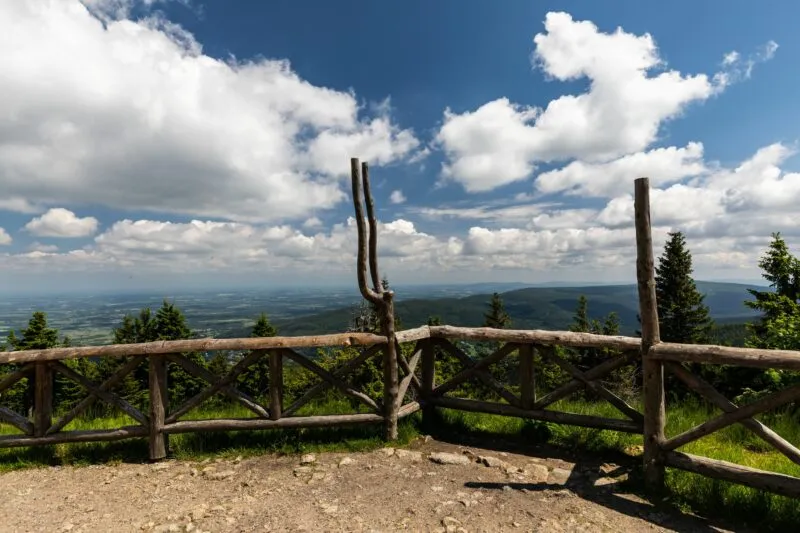 La terrazza panoramica in legno sullo Stóg Izerski apre un'ampia vista sulla valle e sulle montagne lontane, visibile tra i tronchi, gli abeti rossi e le nuvole bianche.