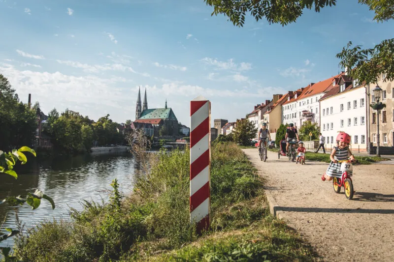Widok rzekę oraz nadbrzeże, po którym jedzie rodzina na rowerach przy słonecznej pogodzie na tle błękitnego nieba.