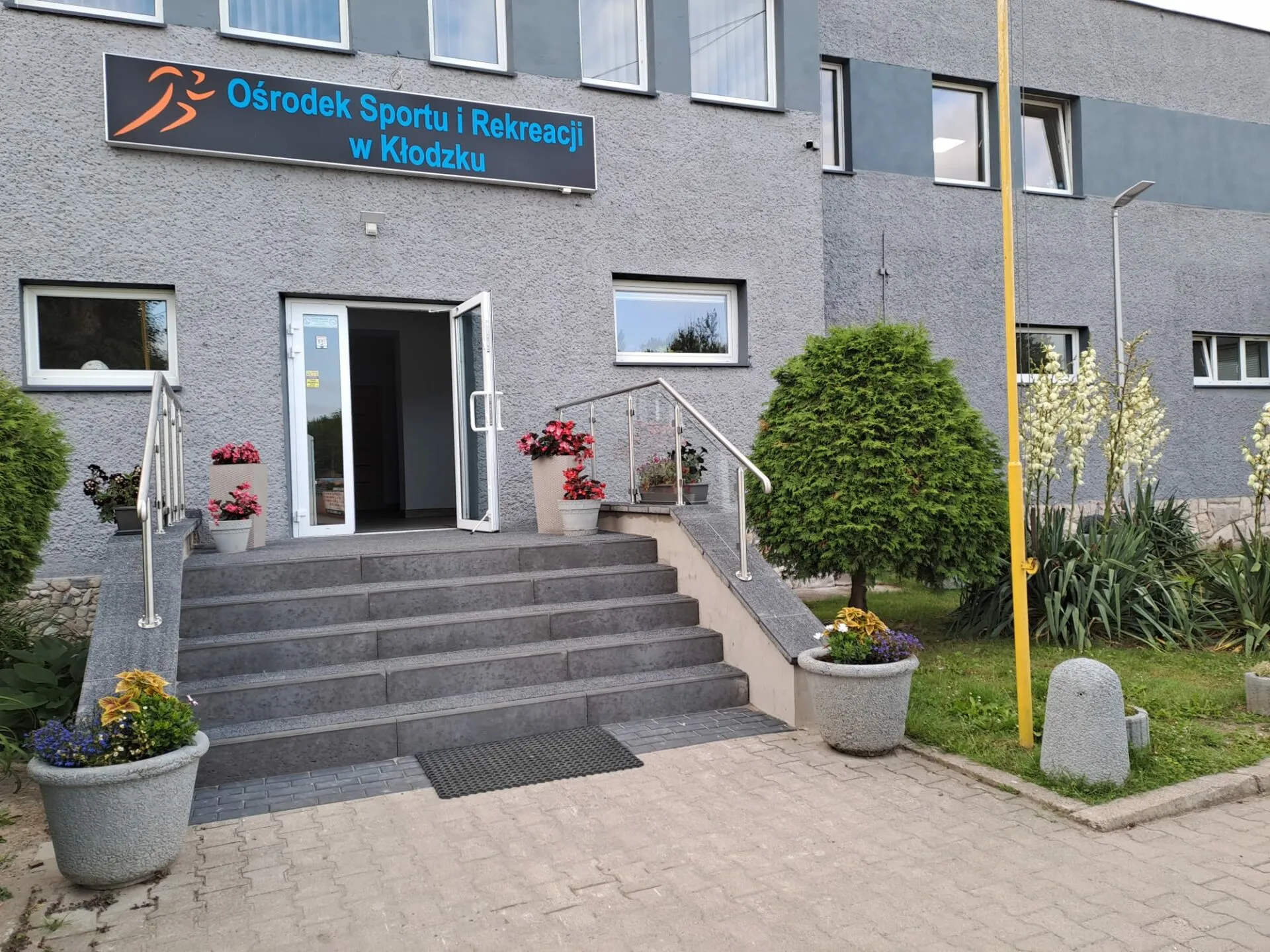 Wejście do szarego, dwupiętrowego budynku, do którego prowadzą schody, zabezpieczone poręczą. Przed wejściem znajdują się donice z nasadzeniami oraz kawałek drobnej ogrodowej zieleni.