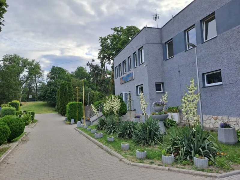 Elewacja szarego dwupiętrowego budynku, do którego prowadzą schody, zabezpieczone poręczą. Przed wejściem znajduje się kawałek drobnej ogrodowej zieleni oraz wybrukowana droga.