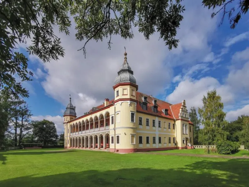 Pałac z jasnożółtą elewacją, czerwonym dachem, wokół pałacu piękny, zadbany trawnik.