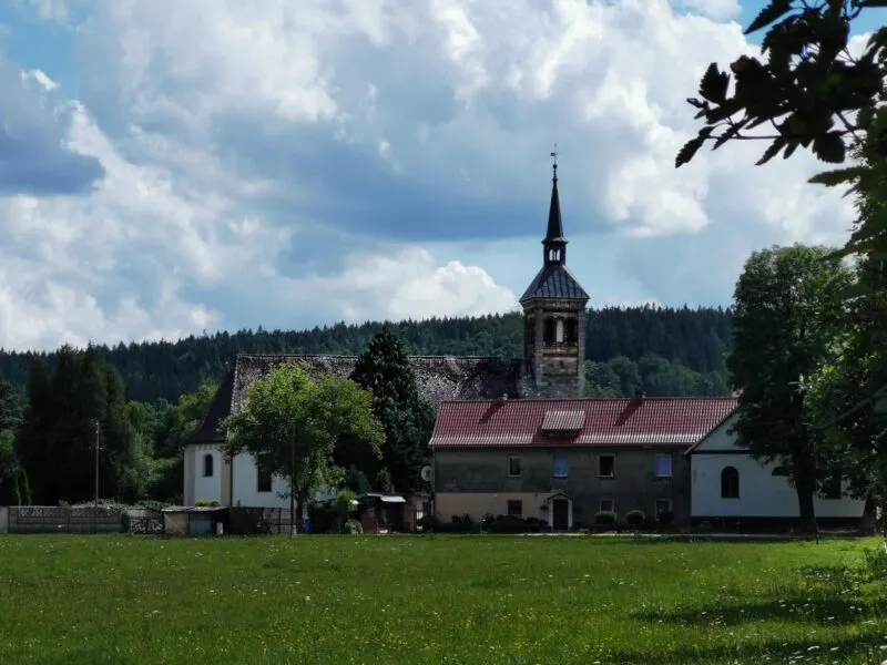 Kamiennny budynek kościoła z jedną wieżą, wśród pól i budynków gospodarczych.