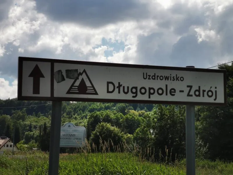 Biały znak drogowy z brązową ramką, wskazujący kierunek do Uzdrowiska Długopole-Zdrój.